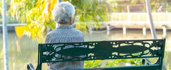 An elderly woman sits on a bench near a large pond, on her own.